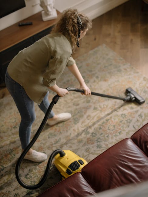 A woman with curly hair wearing a green jacket, blue jeans, and white sneakers is engaged in deep cleaning a residential living room. She is operating a yellow and black vacuum cleaner with a long hose, focusing on a patterned, cream-colored rug that covers a wooden floor. The room has a wooden sideboard against the wall with a white decorative item and a remote control placed on top. The lighting is warm and natural, highlighting the cleanliness of the space. Carpet Cleaning Harrow specializes in surface cleaning and sanitisation, ensuring hygienic and well-maintained environments, as demonstrated by the detailed cleaning process in this domestic setting.
