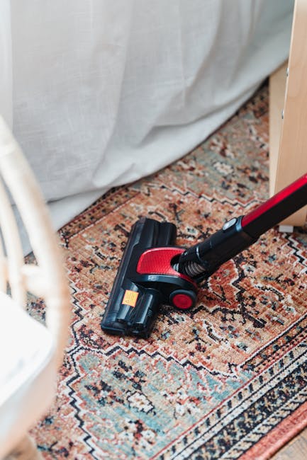 A handheld vacuum cleaner with a red and black design is positioned on a patterned Persian-style rug, which features intricate geometric and floral motifs in shades of red, blue, beige, and brown. The rug is laid out on a wooden floor in a well-lit room. Behind the rug, a light-colored fabric curtain hangs partially white, providing a neutral backdrop. The vacuum cleaner appears to be in use or ready for surface cleaning as part of a deep cleaning process. The scene is captured in a domestic setting, highlighting surface cleaning and maintenance of the rug by Carpet Cleaning Harrow, emphasizing hygiene and cleanliness in a residential space.
