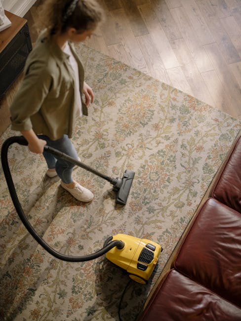 A woman with light skin and brown hair tied back, wearing a light blue striped shirt and blue jeans, is kneeling on a patterned rug in a living room. She is cleaning a wooden floor with a blue cloth,