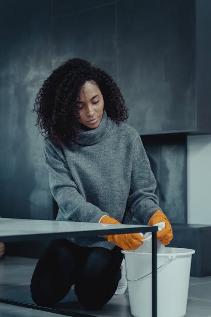 A woman with curly black hair, dressed in a grey sweater and wearing orange rubber gloves, is kneeling on the floor in a modern kitchen or commercial space with dark, polished surfaces and a sleek, minimalist design. She is handling a small cleaning cloth, positioned over a white plastic bucket, and appears to be engaged in surface cleaning or sanitisation. The environment features reflective countertops, possibly made of stainless steel or ceramic, with clean and dust-free surfaces illuminated by ambient lighting. Visible behind her are dark cabinetry and a wall with a smooth, matte finish, emphasizing a professional cleaning setting, consistent with services offered by Carpet Cleaning Harrow under their same-day emergency stain response for domestic or commercial cleaning. The space is tuned for hygiene and thorough cleaning processes, aligning with the focus on deep cleaning and surface sanitisation.
