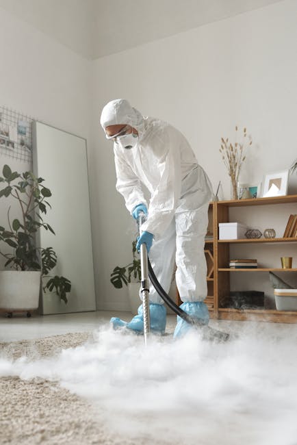 A woman with curly black hair, dressed in a grey sweater and wearing orange rubber gloves, is kneeling on the floor in a modern kitchen or commercial space with dark, polished surfaces and a sleek, minimalist design. She is handling a small cleaning cloth, positioned over a white plastic bucket, and appears to be engaged in surface cleaning or sanitisation. The environment features reflective countertops, possibly made of stainless steel or ceramic, with clean and dust-free surfaces illuminated by ambient lighting. Visible behind her are dark cabinetry and a wall with a smooth, matte finish, emphasizing a professional cleaning setting, consistent with services offered by Carpet Cleaning Harrow under their same-day emergency stain response for domestic or commercial cleaning. The space is tuned for hygiene and thorough cleaning processes, aligning with the focus on deep cleaning and surface sanitisation.