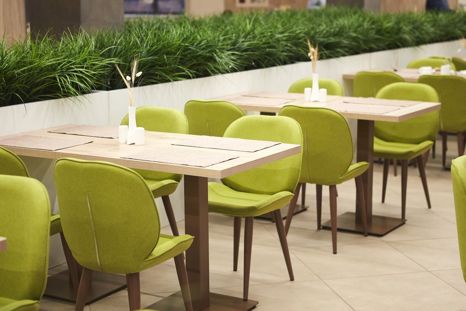 Inside a modern boutique cafe, there are multiple light wooden tables with beige table runners, each adorned with small white vases containing dried floral arrangements. Bright green upholstered chair